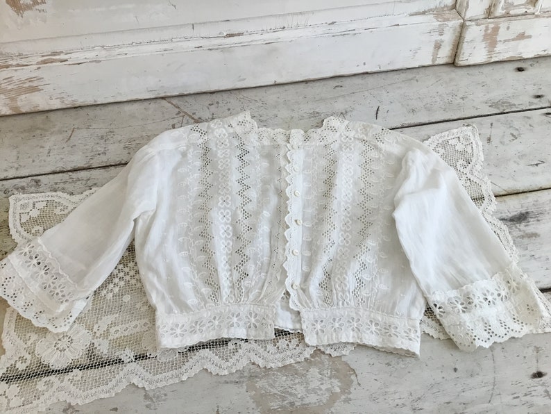 May include: A white, vintage-style blouse with long sleeves and intricate lace detailing. The blouse features a button-up back and delicate floral and geometric patterns. The sleeves and hem are trimmed with scalloped lace.