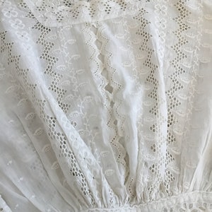 May include: A white, eyelet embroidered blouse with intricate floral and leaf patterns. The blouse features a high, ruffled neckline and a gathered waist. The fabric is delicate and semi-sheer, with detailed cutwork throughout the design.