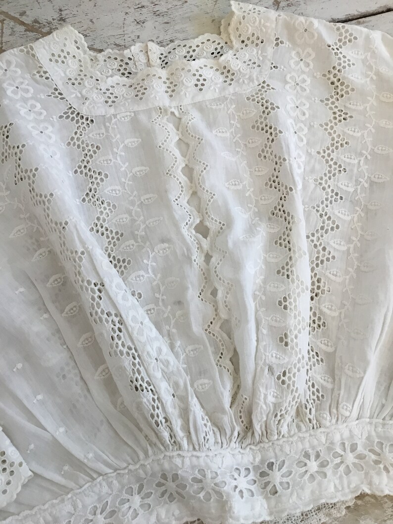 May include: A white, eyelet embroidered blouse with intricate floral and leaf patterns. The blouse features a high, ruffled neckline and a gathered waist. The fabric is delicate and semi-sheer, with detailed cutwork throughout the design.