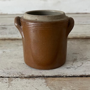 May include: A small, brown ceramic jar with two side handles. The jar has a light-colored rim and a dark brown body. The jar is cylindrical in shape and appears to be vintage or antique.