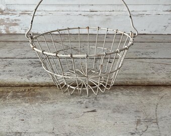 May include: A vintage-style, white wire basket with a handle. The basket has a rounded shape and is made of thin metal wires. The handle arches over the top. The basket sits on a weathered, light-colored wooden surface.