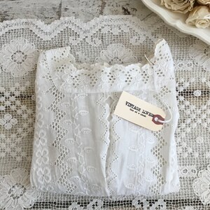 May include: A folded white eyelet fabric garment with a tag that reads "VINTAGE LOVES One of a kind." The garment is placed on a white lace doily. Dried roses are in a white dish in the background.