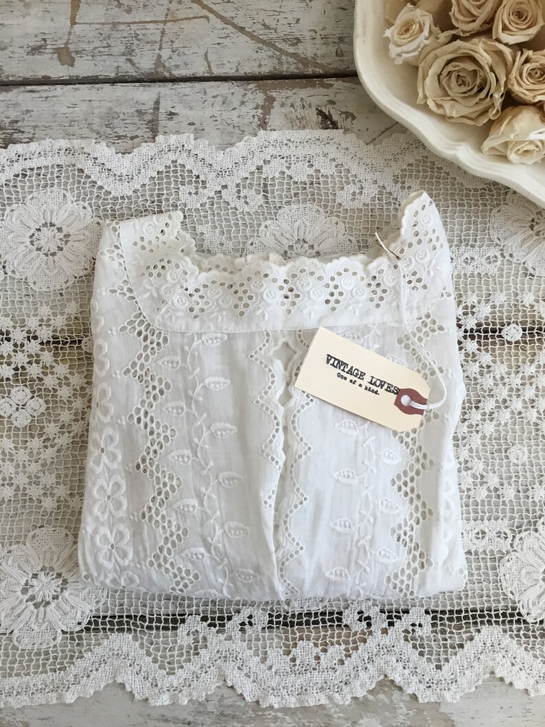 May include: A folded white eyelet fabric garment with a tag that reads "VINTAGE LOVES One of a kind." The garment is placed on a white lace doily. Dried roses are in a white dish in the background.