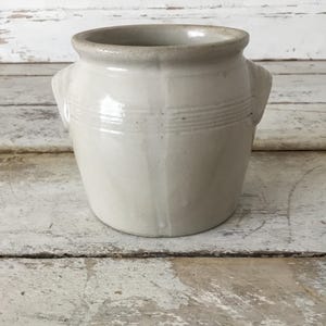 May include: A cream-colored, antique-style ceramic pot with two small handles. The pot has a wide, slightly flared rim and horizontal etched lines around the middle. The pot is sitting on a distressed, white-painted wooden surface.
