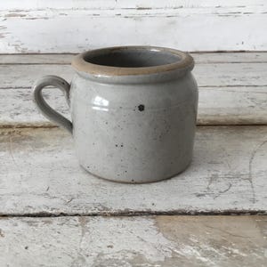 May include: A small, speckled gray ceramic mug with a rounded body and a curved handle. The rim is a lighter tan color. The mug sits on a weathered, white-painted wooden surface. The mug is likely for beverages.