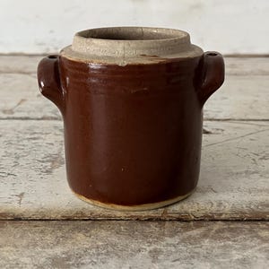 May include: A small, brown ceramic crock with a light-colored rim and two small handles. The crock has a cylindrical shape and a slightly textured surface. It is likely an antique or vintage kitchenware item.