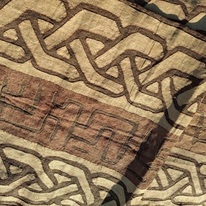 Puede incluir: Primer plano de un textil con un patrón geométrico repetitivo en tonos marrones y beige. El diseño presenta formas y líneas entrelazadas, creando una apariencia texturizada. La tela tiene una estética natural y terrosa.