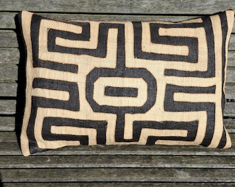 May include: Rectangular decorative pillow with a geometric pattern. The pillow features a beige background with a repeating black maze-like design. The pillow is made of a textured fabric and is placed on a wooden surface.