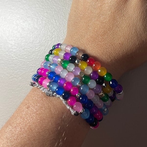 May include: A collection of colorful beaded bracelets in various shades, including pink, blue, green, yellow, and purple. The bracelets are made of small, round beads and are strung together with a silver-colored cord. The bracelets are stacked on top of each other.