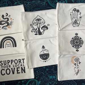 May include: A collection of white canvas zipper pouches with various black and gold mystical designs. Designs include a snake, mushroom, hand with eye, mandala, potion bottle, rainbow, rose, and the text "SUPPORT YOUR LOCAL COVEN".