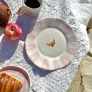 Könnte beinhalten: Ein weißer Keramikteller mit rosa Streifen und einem goldenen Croissant-Design. Eine Tasse Kaffee mit blauen Streifen und ein roter Apfel sind ebenfalls zu sehen. Der Teller gehört zu einem Frühstück oder Brunch.