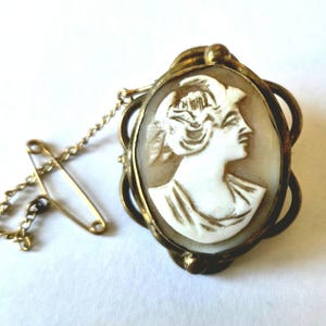 May include: Vintage cameo brooch with a white profile of a woman in an oval, gold-toned frame. A safety pin and chain are attached. A classic piece of jewellery.