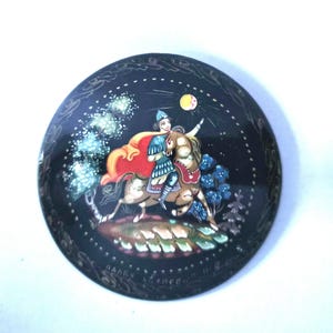 May include: A round black lacquer box lid featuring a hand-painted scene of a figure in blue armour and a red cape riding a brown horse. The scene includes a yellow moon, white trees, and blue foliage. Cyrillic text is visible at the bottom edge.