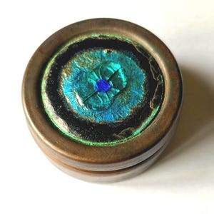 May include: A small, round box with a copper-toned exterior. The lid has a central design of layered teal and blue, reminiscent of a peacock's eye, framed by black and green. This decorative box is likely for storing small items, such as jewelry.