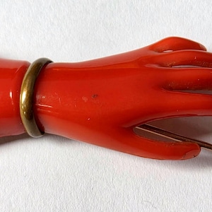 May include: A vibrant red hand-shaped brooch with a gold-colored band around the wrist. The brooch has a pin on the back for attachment. The hand is in a relaxed pose, with fingers slightly curved.