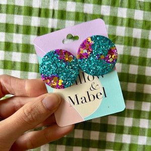 May include: Round teal glitter stud earrings with pink, gold, and purple accents. The earrings are on a light purple and teal card with "Willow & Mabel" printed on it. The earrings are a fun accessory.