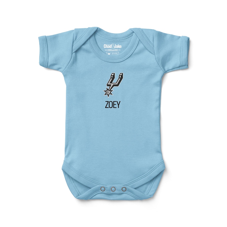 May include: A light blue baby onesie with a black and white graphic of a spur and star, with the name "ZOEY" printed below. The onesie has short sleeves and three snap closures at the bottom.