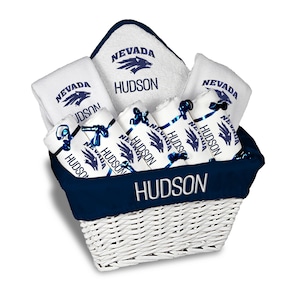May include: A white wicker basket filled with white towels and washcloths, all bearing the text "NEVADA HUDSON" in navy blue. The basket has a navy blue trim and is tied with navy blue ribbons. The towels and washcloths are neatly arranged.