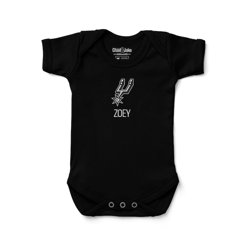 May include: Black baby onesie with a white graphic of a spur and starburst design, with the name "ZOEY" below. The onesie has short sleeves and three snaps at the bottom. The brand name "Chad & Jake" is printed at the top.