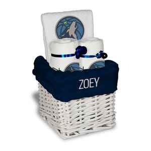 May include: A white wicker basket filled with a navy blue blanket, rolled white towels, and a larger white towel. The towels have a wolf logo and are tied with blue ribbons. The blanket is embroidered with the name "ZOEY".