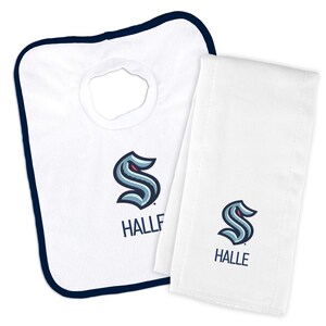 May include: White baby bib and burp cloth set. The bib has a navy blue trim and a neck opening. Both items feature a stylized blue and teal logo with the word "HALLE" in navy blue.