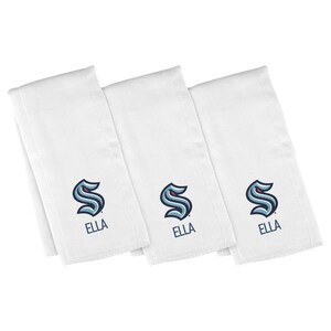 May include: Three white cloth napkins with the Seattle Kraken logo embroidered in navy blue and teal. The name "ELLA" is embroidered below the logo in navy blue. The napkins are folded and stacked.