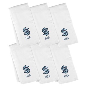 May include: Seven white cloth napkins with a navy blue and teal logo and the name "ELLA" embroidered on each. The logo is a stylized letter "S" with a wave design. The napkins are folded and stacked.