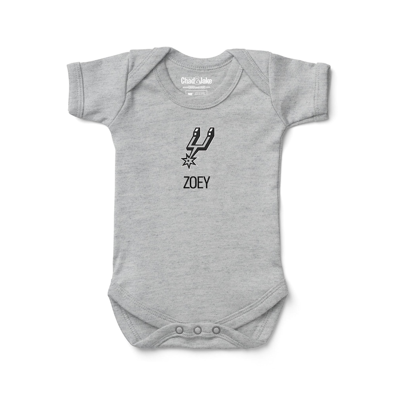 May include: A gray baby bodysuit with short sleeves. The bodysuit features a black graphic of a spur and star, with the name "ZOEY" printed below. The brand "Chad & Jake" is printed on the neckline.