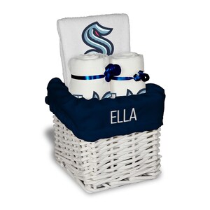 May include: A white wicker basket holds a navy blue blanket embroidered with the name "ELLA", and rolled white towels. The towels are decorated with a navy blue and teal logo and tied with navy blue ribbons. A folded white towel with the same logo is in the back.