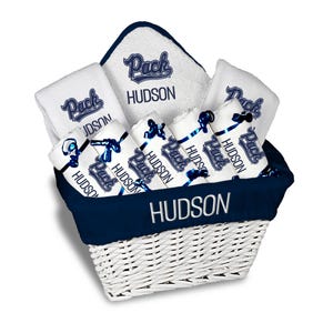 May include: A white wicker basket filled with white towels and washcloths, all bearing the text "Pack HUDSON" in navy blue. Rolled towels are tied with navy blue ribbons. The basket's rim is navy blue, with "HUDSON" embroidered in white.