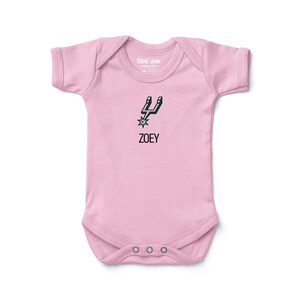 May include: A light pink baby onesie with a black graphic of a stylized spur and starburst, with the name "ZOEY" below. The onesie has short sleeves and three snaps at the bottom.