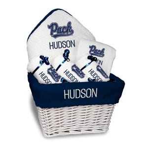 May include: A white wicker basket filled with white and navy blue towels. The towels are embroidered with the word "Pack" and the name "Hudson". The basket has a navy blue lining and trim.