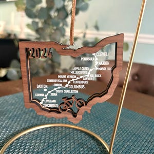 May include: A wooden ornament shaped like the state of Ohio with a bicycle route marked on it. The ornament is brown and has a clear acrylic insert with the year 2024 and the names of cities and towns in Ohio printed on it.