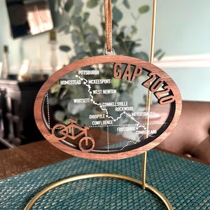 May include: A wooden Christmas ornament with a clear acrylic insert featuring a map of the Great Allegheny Passage bike trail. The map includes the names of towns along the trail, including Pittsburgh, Homestead, McKeesport, West Newton, Connellsville, Rockwood, Meyersdale, Ohiopyle, Confluence, Frostburg, and Cumberland. The ornament also features a silhouette of a bicycle.
