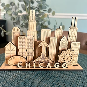 May include: Wooden Chicago skyline model featuring iconic buildings and landmarks. The laser-cut design includes the city's name at the base. The model is made of light-colored wood and is displayed on a blue surface.