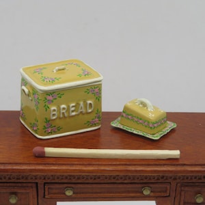 May include: A miniature yellow bread bin and butter dish with floral patterns are on a wooden table. The bread bin has the word "BREAD" in white. A large match is also on the table.