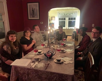May include: A group of people are sitting around a table set for a dinner party. The table is covered in a grey tablecloth with a floral pattern. There are plates, glasses, and silverware set on the table. The people are all smiling and looking at the camera.