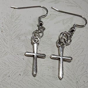 May include: A pair of silver-toned dangle earrings featuring a simple cross charm suspended from a standard fishhook ear wire. The cross pendants have a smooth, polished metallic finish and are attached to the ear wires with small circular jump rings.