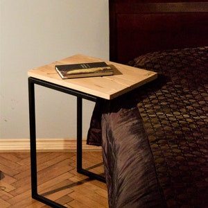 Könnte beinhalten: Ein kleiner, moderner Beistelltisch mit einer hellen Holzplatte und einem schwarzen Metallrahmen. Ein Buch mit dunklem Einband liegt auf dem Tisch. Der Tisch steht neben einem Bett mit einer gemusterten Bettdecke.