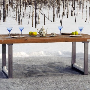 Può includere: Un tavolo rustico in legno con gambe in metallo in un paesaggio innevato. Il tavolo è apparecchiato con bicchieri blu, piatti floreali, una teiera, una tazza da tè e mele verdi. La scena evoca un senso di cena all'aperto.