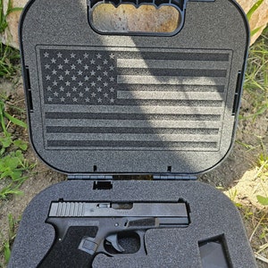 May include: A black handgun with a silver slide is inside a black hard case with a gray foam interior. The case has a handle and a US flag design on the lid.