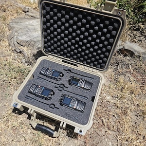 May include: An open, tan-colored hard case containing four black two-way radios. The radios are neatly arranged in a foam insert. The case has a black handle and is sitting on a natural outdoor surface.