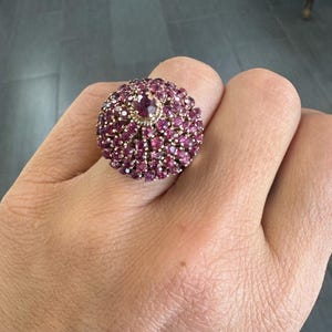 May include: A large, round ring with a cluster of small, deep pink gemstones. The ring's center features a larger, similar-colored stone. The ring is set on a gold-colored band.