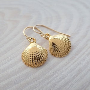 May include: A pair of gold-toned drop earrings featuring textured seashell charms. Each earring consists of a small, detailed scallop shell pendant suspended from a simple curved ear wire hook, showcasing a metallic finish and ridges.