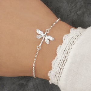 Sterling silver dragonfly bracelet for women with a dainty, detailed nature-inspired dragonfly charm on a delicate adjustable chain. Worn on a woman's wrist with a boho style sleeve for scale.