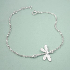 May include: A silver chain bracelet with a delicate dragonfly charm. The dragonfly is detailed with textured wings and a slender body.