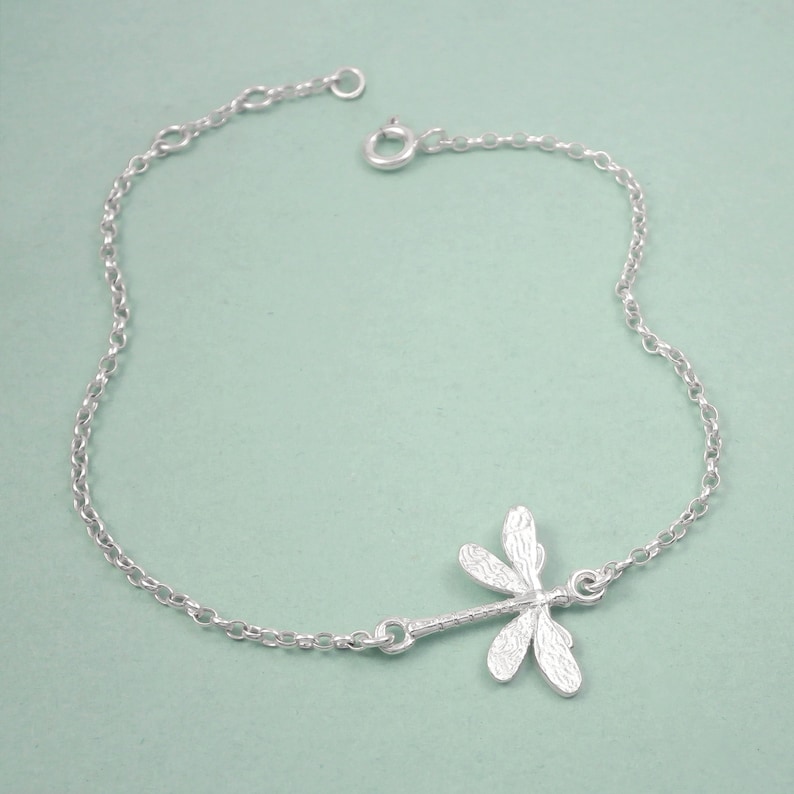 May include: A silver chain bracelet with a delicate dragonfly charm. The dragonfly is detailed with textured wings and a slender body.
