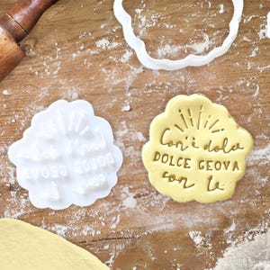 Può includere: Un tagliapasta bianco con il testo "All I need is you" e un biscotto giallo con il testo "Con i dolci dolce Genova con te" in lettere dorate.