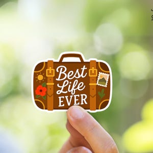 May include: A brown suitcase-shaped sticker with the words "Best Life Ever" in white script. The sticker features a sun, a red flower, and a postage stamp design. The sticker is held by a hand against a blurred green background.