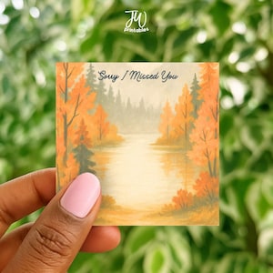 Pode incluir: Um cartão quadrado com uma pintura em aquarela de uma paisagem de outono. A cena inclui um rio, árvores com folhas laranja e amarelas e o texto "Sorry I Missed You". O cartão é segurado por uma mão.